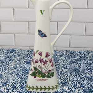May include: A tall white ceramic pitcher with a triangular base, featuring a green letter V near the rim, a blue butterfly, and a botanical illustration of purple Cyclamen Repandum flowers with green leaves and a green vine border at the bottom.