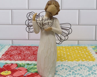 May include: A white porcelain figurine of an angel holding a wire heart that says "Thank you". The angel is standing on a colorful floral patterned fabric.