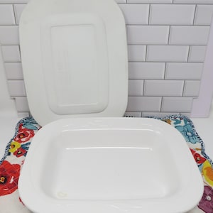 May include: A white rectangular baking dish with rounded corners and a matching lid. The dish sits on a floral patterned cloth. The lid is slightly larger than the dish and has a recessed rectangular area.