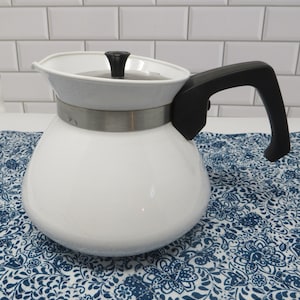 May include: A white teapot with a rounded body, a black handle, and a black knob on the lid. A metallic silver band encircles the upper portion of the teapot, separating the main body from the lid. The teapot features a small spout on the left side.