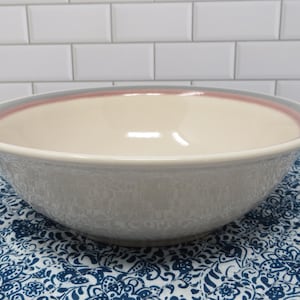 May include: A large, round ceramic bowl with a light gray exterior and a cream-colored interior. The rim features a thin pink band between gray edges. The bowl sits on a blue and white patterned surface.