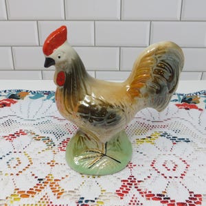 May include: A ceramic rooster figurine with a red comb and wattle, brown and tan body, and a green base. The rooster has a detailed tail and is posed standing upright. The figurine is on a white lace tablecloth with a floral pattern.