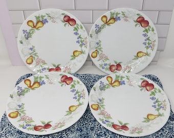 May include: A set of four white ceramic dinner plates, each featuring a circular border pattern of red apples, yellow pears, blue berries, and green leaves. The plates have a smooth, glossy finish and a simple, round shape.