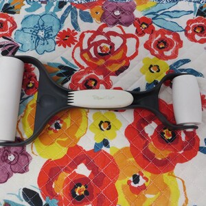 May include: A black and white pastry roller with two white rollers and a white handle. The roller is designed for even pressure when rolling out dough. The background is a floral pattern with red, yellow, and blue flowers.