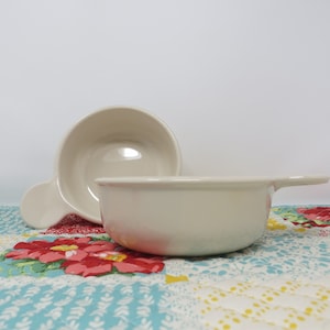 May include: Two off-white ceramic bowls with handles. The bowls are on a colorful patchwork quilt.