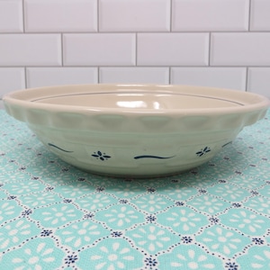 May include: Off-white ceramic pie dish with a scalloped edge and blue floral and line details. The dish sits on a turquoise and white patterned surface, with a white tile background.
