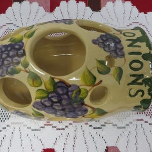 May include: A ceramic onion keeper with a light yellow base, featuring hand-painted purple grapes and green leaves. The word "ONIONS" is written vertically in green. The keeper has multiple circular openings.