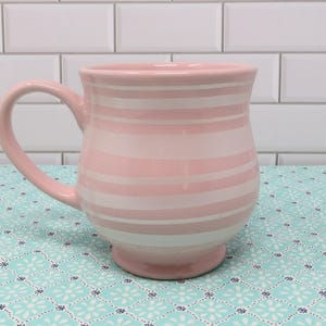 May include: A pink ceramic mug with white stripes. The mug has a wide base and a rounded handle.