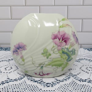 May include: Cream-colored, round ceramic vase with a floral design. The vase features pink and purple morning glory flowers with green leaves and vines. The flowers are painted on the surface of the vase.