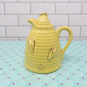 May include: Yellow ceramic honey pot with a beehive design and three gold bees. The pot has a lid and a handle.
