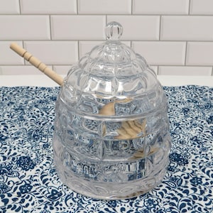 May include: A clear, faceted glass honey pot with a wooden honey dipper. The pot has a domed lid with a small knob. The honey pot is sitting on a blue and white patterned tablecloth. The background is a white brick wall.