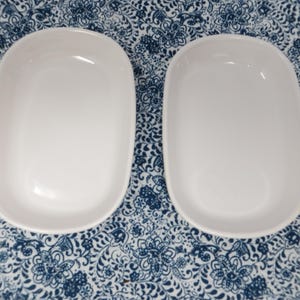 May include: Two identical white oval-shaped ceramic dishes with smooth, rounded edges and a deep basin. The dishes feature a clean, glossy finish and a simple, functional design, presented side-by-side as a matching pair for serving or display.