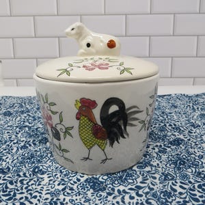 May include: A white ceramic canister with a lid featuring a sculpted cow handle. The canister is decorated with a colorful rooster illustration, green vines, and pink floral patterns. The rooster has a red comb, yellow body, and black tail.