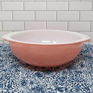 May include: A round, solid pink mixing bowl with a smooth, glossy finish and integrated side handles. The interior of the bowl is white, contrasting with the exterior pink color. The bowl features a wide, open top and a curved, deep basin.