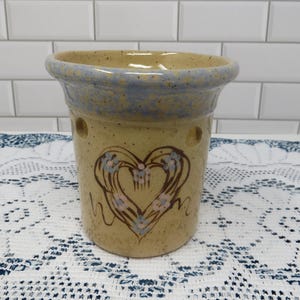 May include: A tan ceramic candle holder with a blue rim and a heart design. The heart is outlined in brown with blue and pink floral accents. The candle holder has two small holes and is sitting on a white lace tablecloth.
