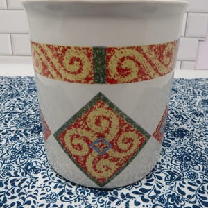 May include: White ceramic container with a wide rim, featuring a decorative band of red and yellow swirls. A diamond pattern with similar colors and blue accents adorns the body. The container sits on a blue and white patterned fabric.