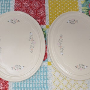 May include: Two round, cream-colored ceramic serving plates with a floral design in shades of pink and gray.