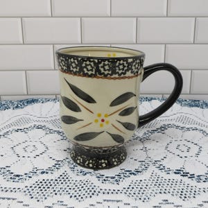 May include: Cream-colored ceramic mug with a black handle and base. The mug features a floral design with black leaves and yellow and red accents. The rim has a decorative black pattern.