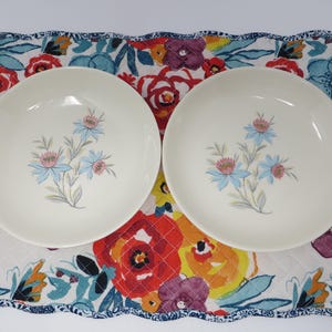 May include: Two off-white ceramic bowls with a floral design featuring blue and pink flowers. The bowls rest on a colorful floral placemat with a quilted pattern. The placemat has a blue patterned border.