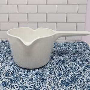 May include: A white ceramic pitcher with a long handle and a heart-shaped spout. The pitcher is sitting on a blue and white floral patterned cloth. The background is a white tiled wall.