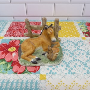 May include: A ceramic figurine of a brown foal lying down next to a small brown and white squirrel. The figurine is set against a backdrop of a wooden fence and a colorful patchwork quilt.