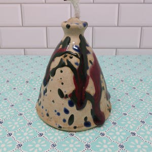 May include: A ceramic oil lamp with a beige base, adorned with dark red and black paint streaks, and blue speckles. The lamp has a flared base and a narrow neck with a wick extending from the top. The lamp is approximately 5 inches tall.