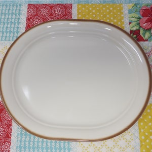 May include: A large, oval-shaped serving platter with a white glaze and a brown rim. The platter is sitting on a colorful patchwork fabric.