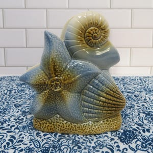 May include: A ceramic sculpture featuring a starfish, seashell, and conch shell in shades of blue, brown, and beige. The starfish has a textured surface, and the conch shell is spiraled. The base is a sandy brown color.