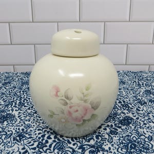 May include: A cream-colored ceramic ginger jar with a matching lid. The jar features a rounded body and a delicate, soft-toned floral design on the front, showcasing pale pink roses and muted green leaves painted in a subtle, watercolor style.
