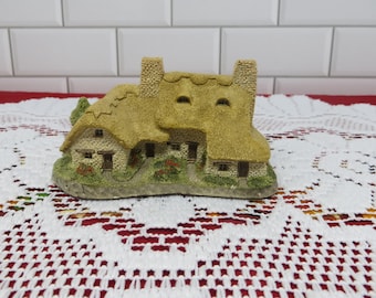 Może przedstawiać: Miniaturowa figurka domku z trzcinowym dachem, dwoma kominami i małymi oknami. Domek wykonany jest z materiału przypominającego kamień i posiada mały ogródek z kwiatami. Figurka stoi na białym, koronkowym obrusie.
