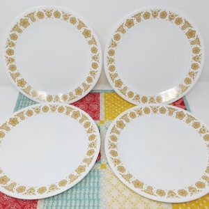 May include: Four white dinner plates with a gold floral pattern border. The plates are arranged on a colorful patterned tablecloth. The floral design is a repeating pattern of stylized flowers and leaves.
