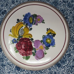 May include: A round ceramic plate with a cream-colored surface featuring a hand-painted floral arrangement in shades of red, yellow, blue, and purple. The plate is bordered by a thin dark red circular line and a wide light gray outer rim.