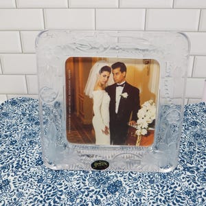 May include: Clear, square photo frame with floral and leaf detailing. The frame holds a wedding photo of a bride and groom. The frame has the word "Studios" at the bottom. The frame is sitting on a blue and white patterned surface.