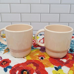 May include: Two matching light peach-colored ceramic mugs with handles. The mugs have a stacked design and are set on a floral patterned quilted surface. The background features white subway tile.