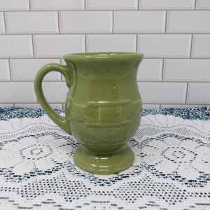 May include: A sage green ceramic mug featuring a rounded body with a raised, embossed basket-weave texture around the center. The mug has a sturdy handle on the left side, a wide rim, and a pedestal base, all finished in a smooth, glossy glaze.