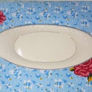 May include: A white oval serving dish with a raised, woven pattern around the rim. The dish is on a blue and white floral tablecloth.