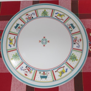 May include: White ceramic Christmas plate with a teal rim and red and green accents. The plate features festive illustrations of Santa Claus, Christmas trees, rocking horses, and houses. A central floral design completes the holiday theme.