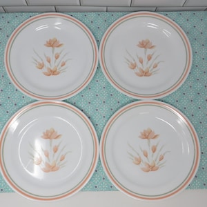 May include: Set of four white dinner plates with a floral design. The plates have a peach colored flower with green leaves and a thin orange and green border.