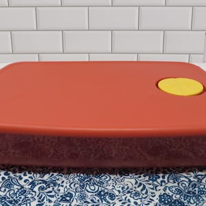 May include: A rectangular, red food storage container with a yellow circular lid. The container sits on a blue and white patterned cloth. The background features a white brick wall.