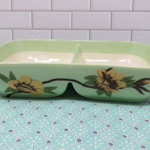 May include: A divided, light green ceramic serving dish with a floral design. The dish has two compartments with a cream-colored interior. Yellow and brown flowers with green leaves decorate the exterior. The dish is on a patterned blue and white surface.