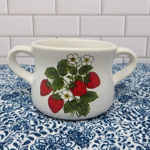 May include: White ceramic mug with two handles, decorated with a strawberry design. The design features red strawberries, green leaves, and white flowers. The mug is sitting on a blue and white patterned surface.
