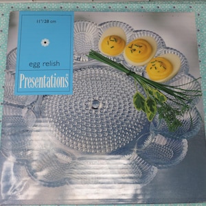 May include: A clear glass serving dish with a textured surface. The dish is 11 inches in diameter and is designed to hold deviled eggs. The dish is surrounded by sprigs of fresh green herbs. The text "egg relish Presentations" is printed on a blue label.