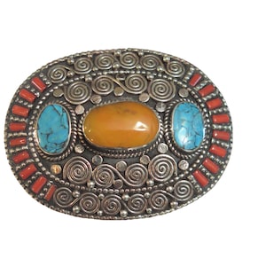 May include: An oval-shaped silver brooch with intricate swirl designs. The brooch features three stones: a large amber-colored stone in the center and two smaller turquoise stones on either side. The brooch is accented with small red coral pieces around the edge.