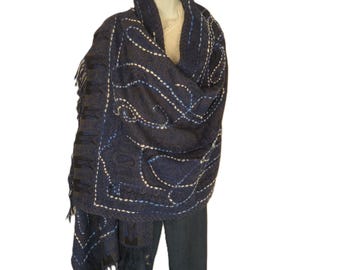 May include: A dark blue shawl with white and blue embroidered details and fringe. The shawl is draped over a mannequin, showcasing the intricate patterns and textures of the fabric. The design features a paisley pattern.
