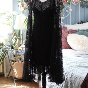 K&ouml;nnte beinhalten: Ein schwarzes Spitzenkleid mit passendem Spitzenumhang wird auf einer Schaufensterpuppe pr&auml;sentiert. Das Kleid hat einen V-Ausschnitt und Spaghettitr&auml;ger. Der Umhang bedeckt die Schultern. Das Kleid ist etwa 102 cm lang.