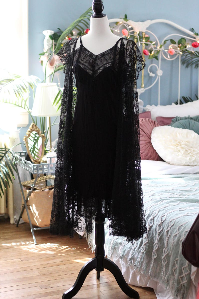 K&ouml;nnte beinhalten: Ein schwarzes Spitzenkleid mit passendem Spitzenumhang wird auf einer Schaufensterpuppe pr&auml;sentiert. Das Kleid hat einen V-Ausschnitt und Spaghettitr&auml;ger. Der Umhang bedeckt die Schultern. Das Kleid ist etwa 102 cm lang.