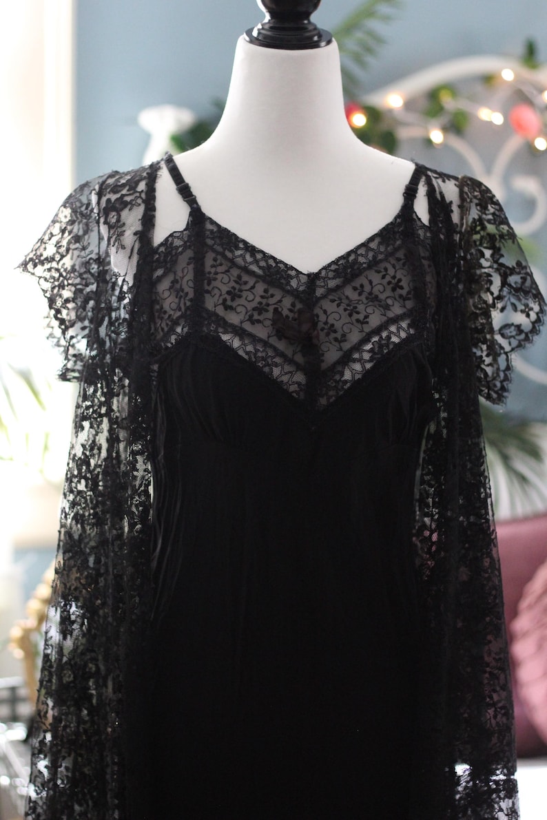 K&ouml;nnte beinhalten: Schwarzes Lingerie-Set im Vintage-Stil auf einer Schaufensterpuppe. Das Set umfasst ein Slip-Kleid mit V-Ausschnitt und zarter Spitze sowie einen passenden Spitzen-Overlay mit kurzen &Auml;rmeln. Das Design ist elegant und romantisch, mit aufwendigen Details.