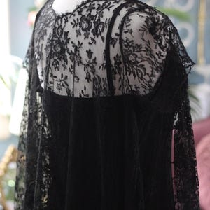 K&ouml;nnte beinhalten: Schwarzes Spitzentop mit floralen Mustern, auf einer Schaufensterpuppe pr&auml;sentiert. Das Top hat eine transparente Spitzenauflage &uuml;ber einem einfarbigen schwarzen Stoff, mit einem gewellten Rand am Ausschnitt und an den &Auml;rmeln. Das detaillierte Spitzendesign verleiht Eleganz.