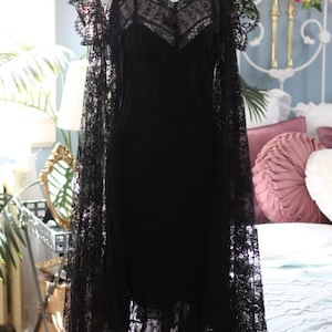 K&ouml;nnte beinhalten: Ein schwarzes Vintage-Kleid mit Spitzen-Overlay, pr&auml;sentiert auf einer Schaufensterpuppe. Das Kleid hat einen V-Ausschnitt und zarte Tr&auml;ger. Die Spitzen&auml;rmel im Cape-Stil verleihen eine elegante Note. Das Kleid ist ein klassisches St&uuml;ck Vintage-Mode.