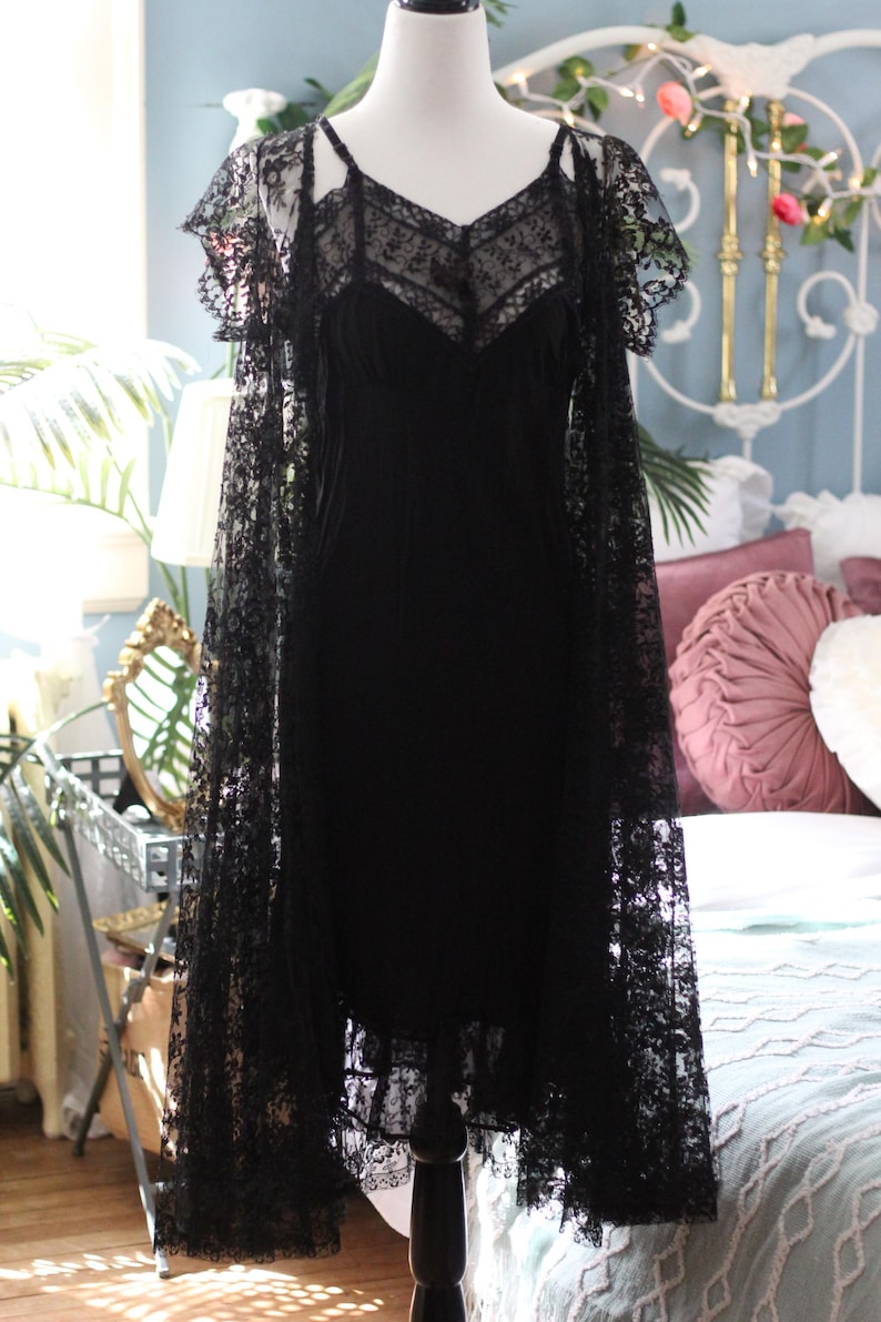 K&ouml;nnte beinhalten: Ein schwarzes Vintage-Kleid mit Spitzen-Overlay, pr&auml;sentiert auf einer Schaufensterpuppe. Das Kleid hat einen V-Ausschnitt und zarte Tr&auml;ger. Die Spitzen&auml;rmel im Cape-Stil verleihen eine elegante Note. Das Kleid ist ein klassisches St&uuml;ck Vintage-Mode.